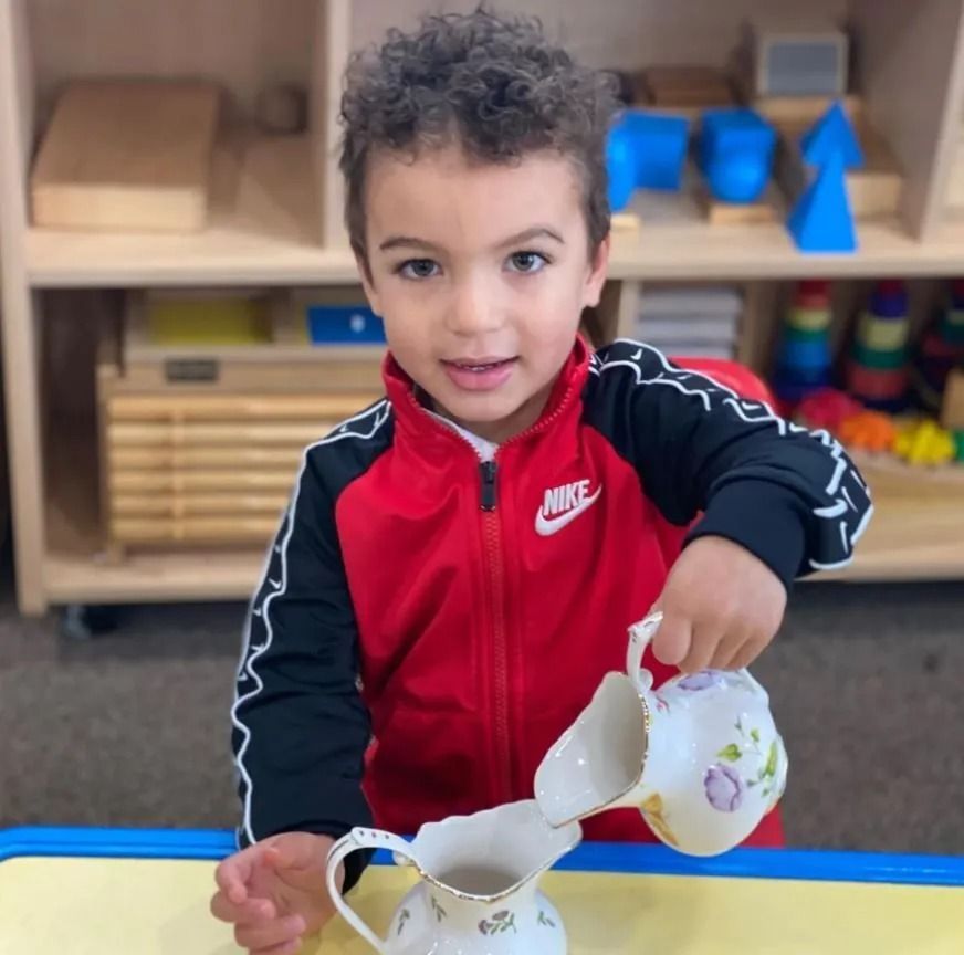 Boy pouring from a small white floral pitcher into another; red jacket, school setting.
