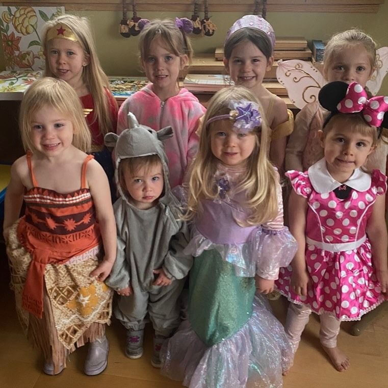 Nine children in costumes pose indoors. They wear outfits like Wonder Woman, Moana, Minnie Mouse, a mermaid, and a shark.