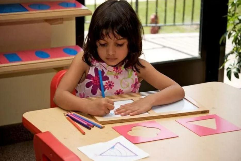 Girl drawing at a desk with shape stencils and colored pencils in a well-lit room.