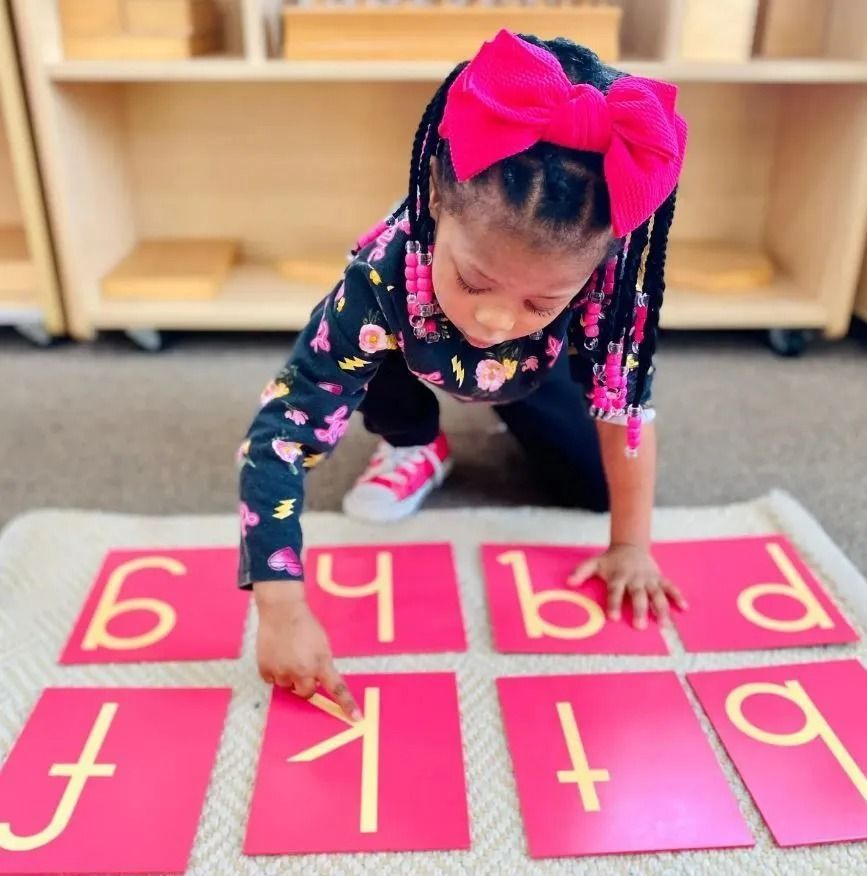 Child with a pink bow plays with letter cards on a rug in a classroom.