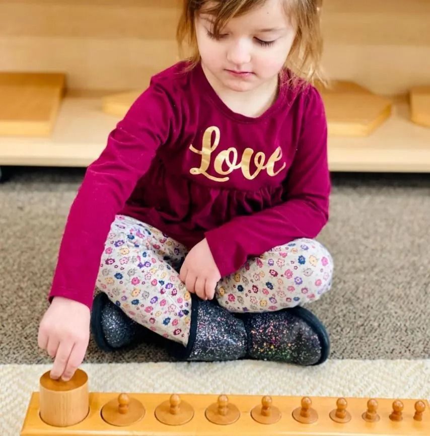 Child seated on the floor playing with a wooden cylinder block set in a classroom setting.