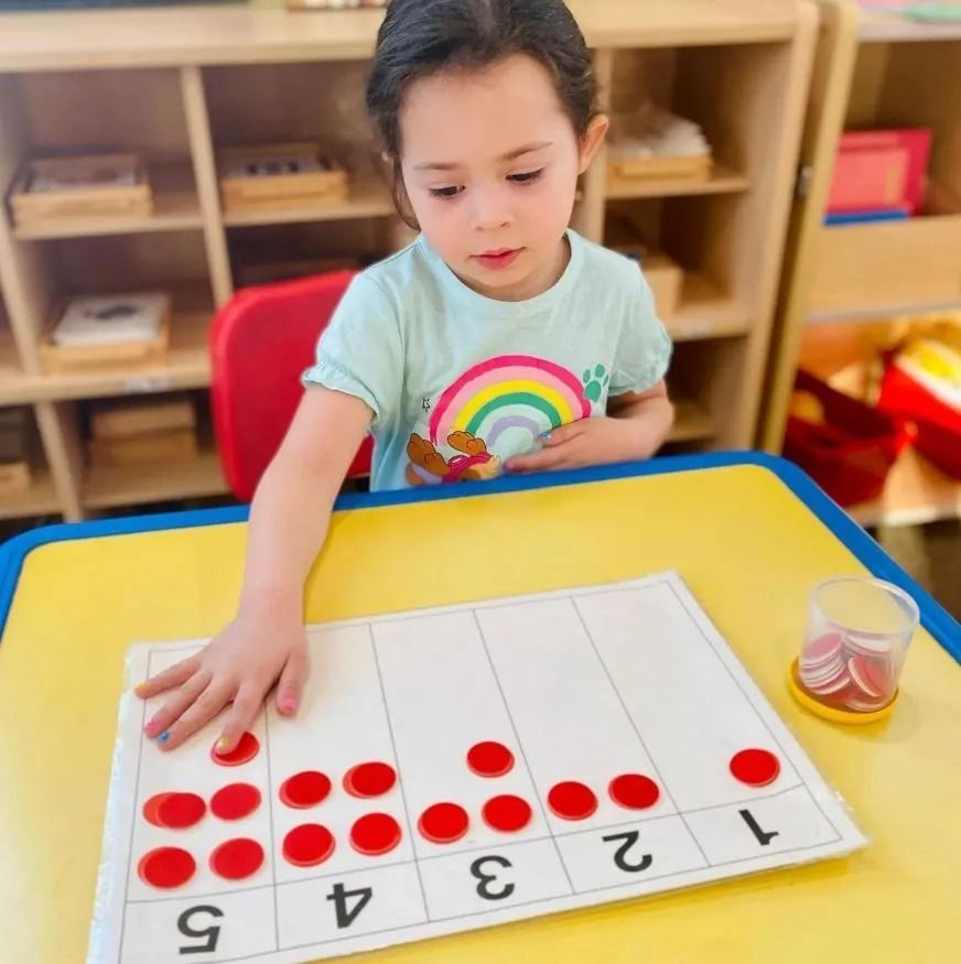 Girl using red counters to learn counting, at a desk with number labels.