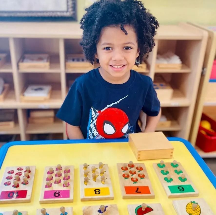 Boy smiling at a table with number learning tools, wooden blocks, and shelves in the background.
