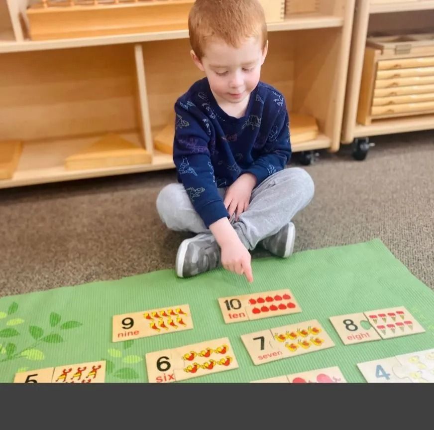 Boy seated on floor points to a math card. Other number cards and wooden shelves are in the background.