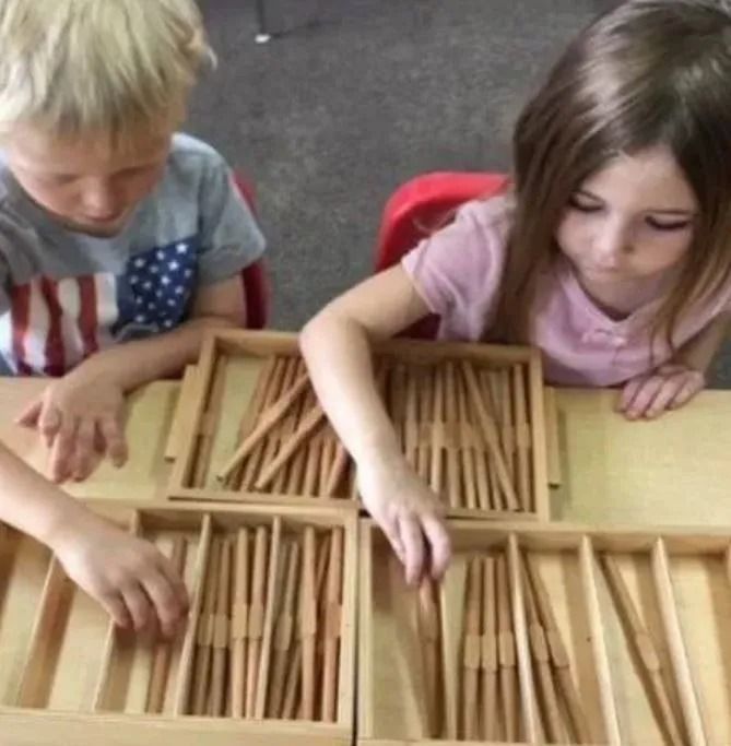 Children using wooden sticks in trays at a table.