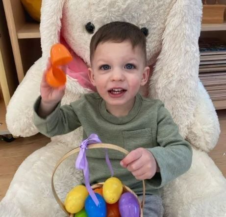 Boy holding an Easter basket with colorful eggs, orange eggshell, smiling in front of a giant stuffed bunny.