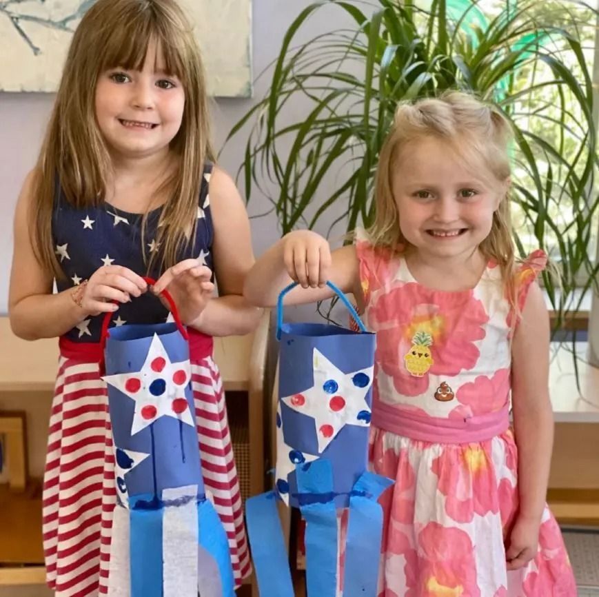 Two girls smiling, holding red, white, and blue paper lanterns, decorated with stars, indoors.