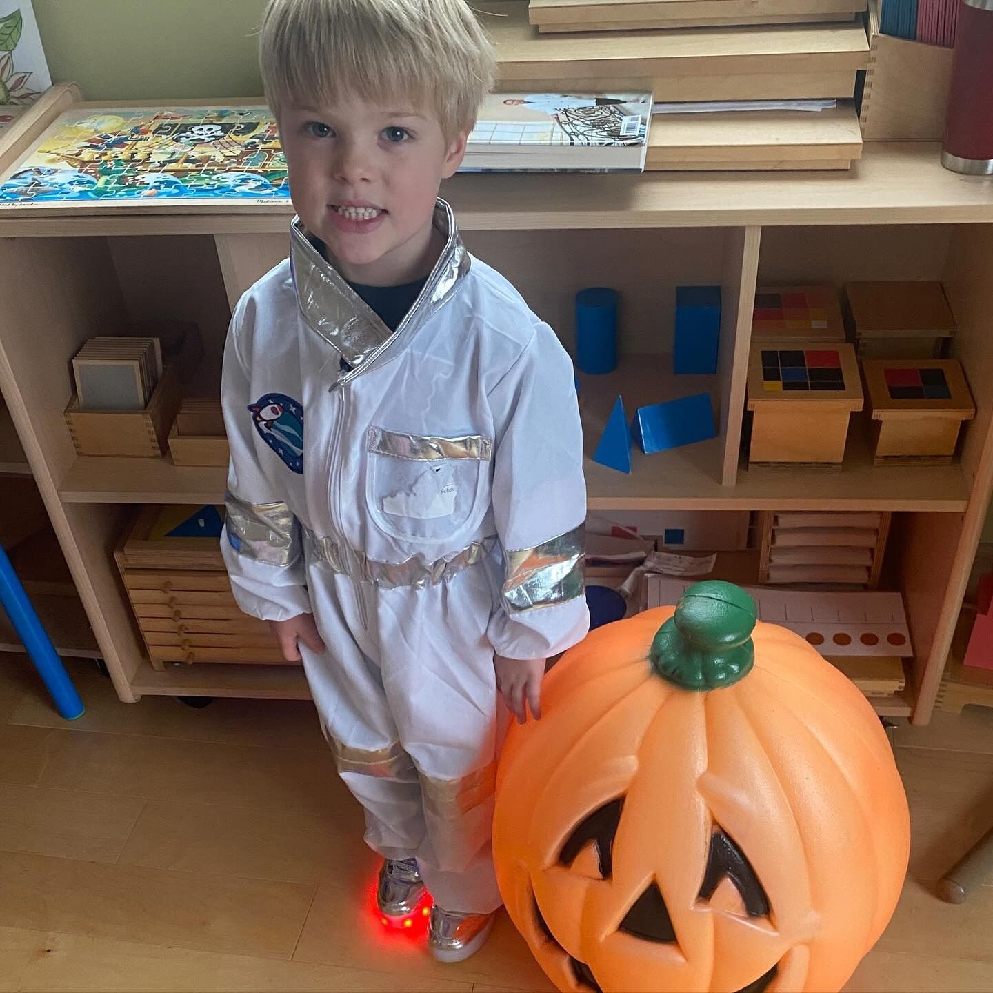 Boy in astronaut costume with lit-up shoes, stands next to a lit orange pumpkin, in a room with shelves.