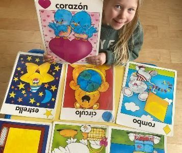 Girl holding a Spanish flashcard with blue birds on a heart, surrounded by shape flashcards on a table.