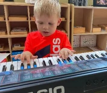 Boy in a red shirt playing a keyboard in front of wooden shelves.