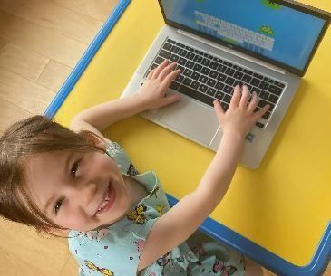 Child smiling, typing on a laptop at a yellow table.