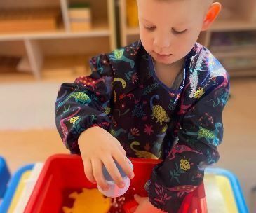 Child playing with water in a red bin, wearing a dinosaur print smock.