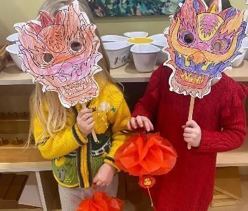 Two children with dragon masks and paper lanterns, indoors. One child wears a yellow sweater, the other a red dress.