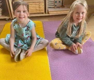 Two young girls smiling, sitting cross-legged on yoga mats; one yellow, one purple.