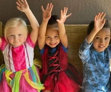 Three smiling children in costumes raise their arms.