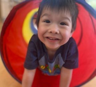 Smiling child in a blue shirt crawls out of a colorful play tunnel.
