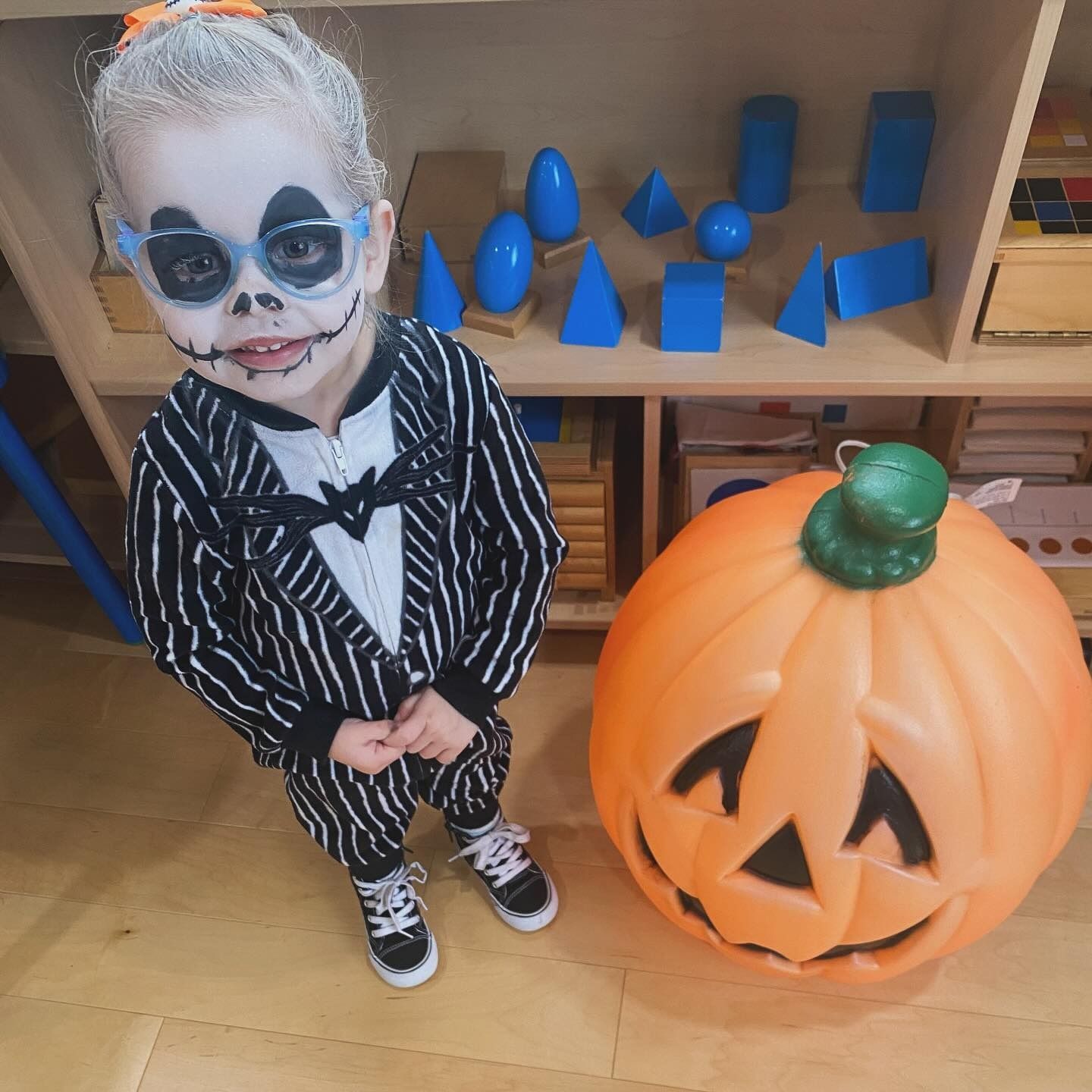 Child in Jack Skellington costume poses with a large pumpkin prop.