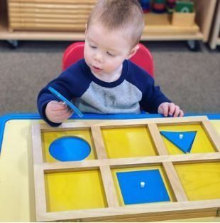 Child playing with a wooden puzzle board with blue and yellow shapes, at a table.