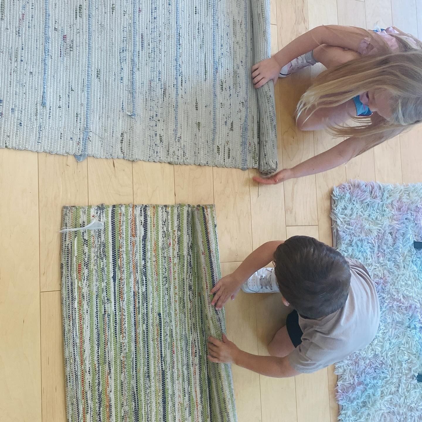 Two children rolling out colorful woven rugs on a wooden floor.