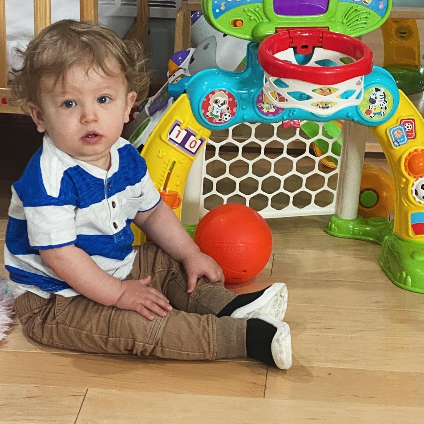 Child sitting with a toy basketball set, orange ball, wearing blue and white striped shirt and brown pants.