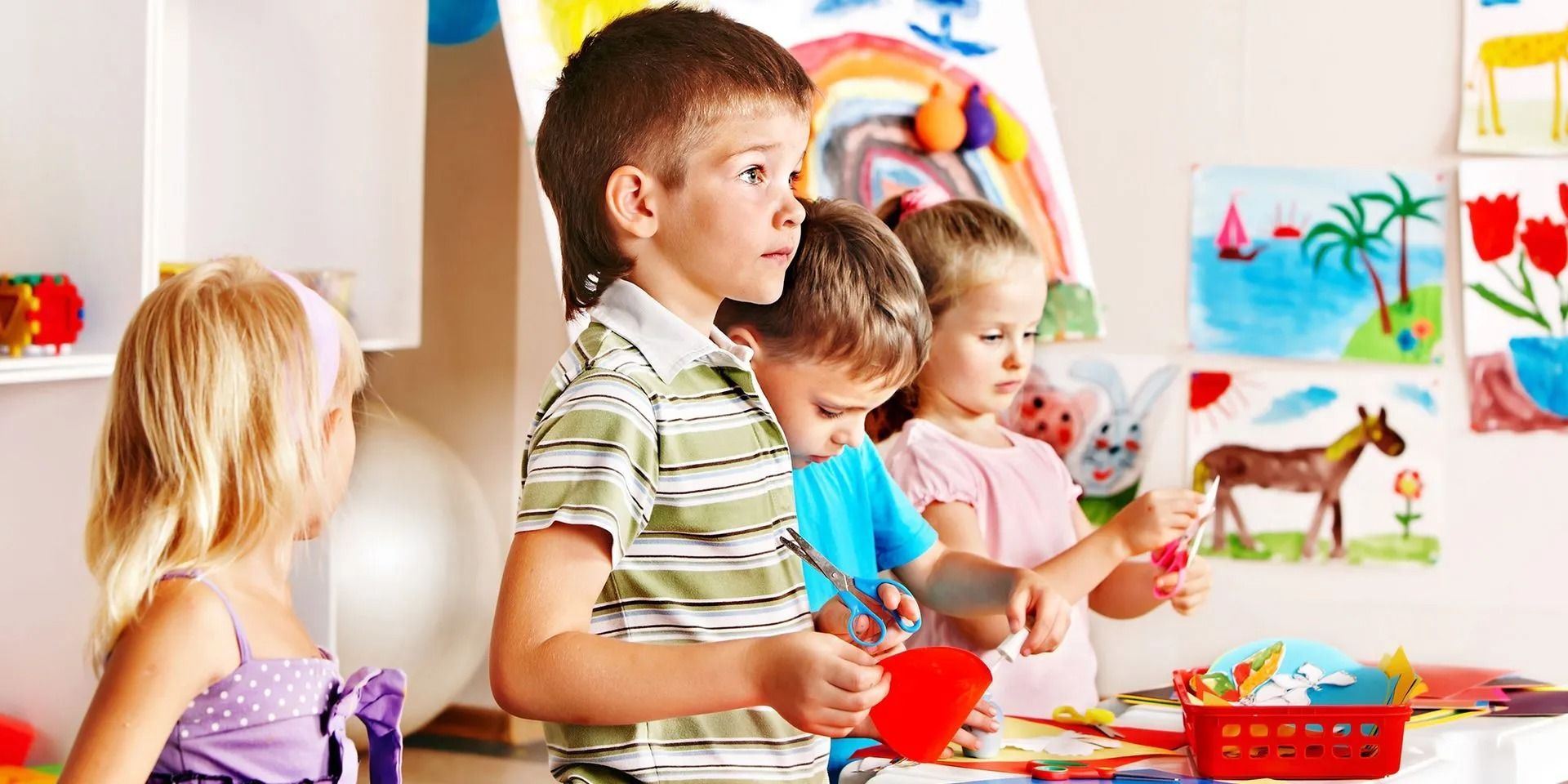 Children painting at a table in a classroom, with drawings on the wall and art supplies nearby.