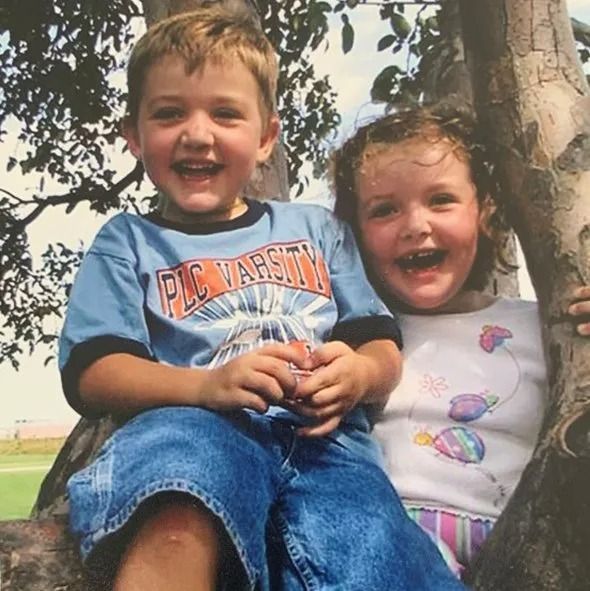 Two children smiling widely, sitting in a tree. The boy wears a blue shirt and jeans; the girl, a white shirt and patterned skirt.