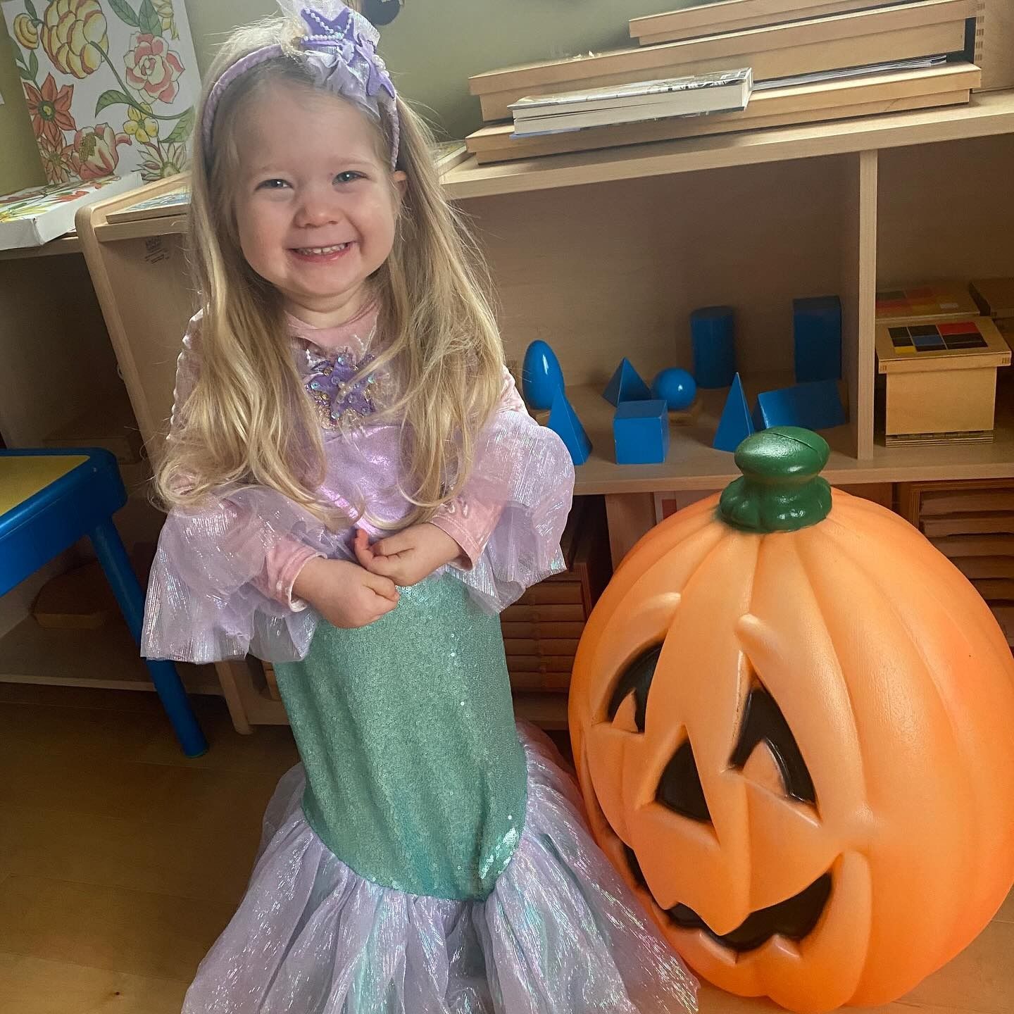Child dressed as a mermaid smiles next to a large orange pumpkin.