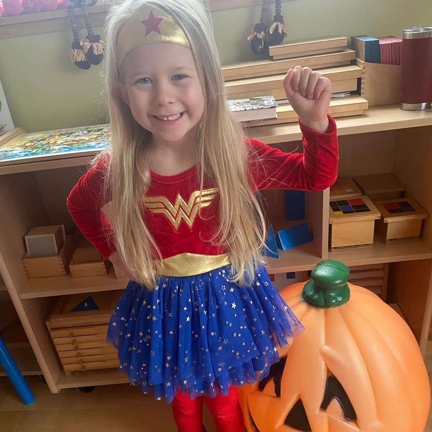 Girl in Wonder Woman costume, smiling, raising fist, next to a pumpkin.