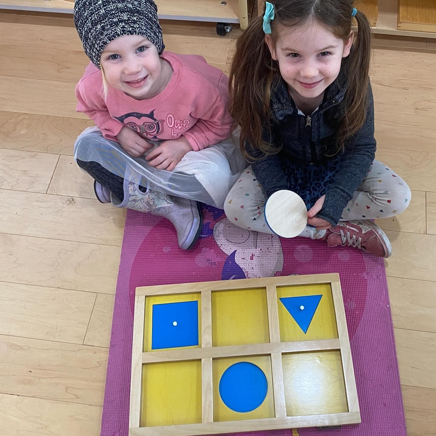 Two children smiling, seated with a wooden shape matching puzzle on a mat.