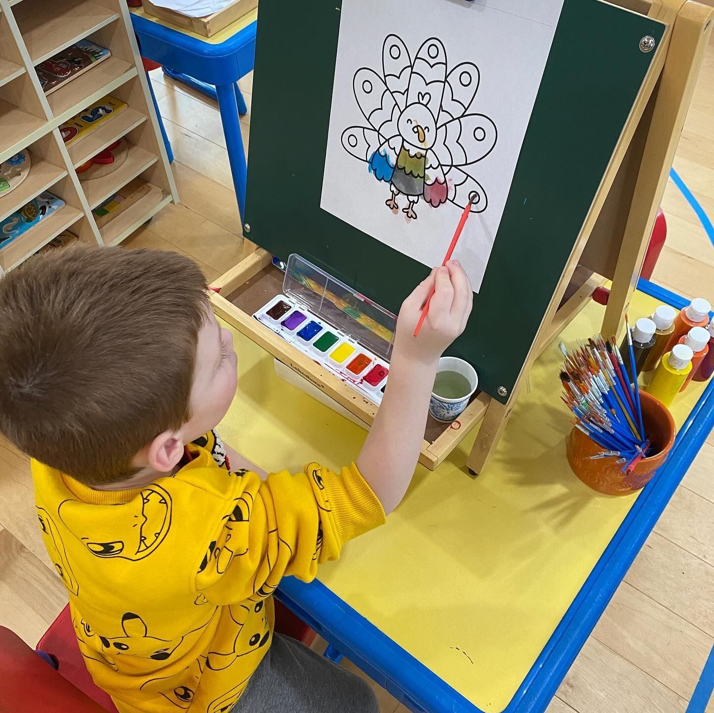 Child painting a turkey on an easel. Yellow table and shirt. Paint palette and brush visible.