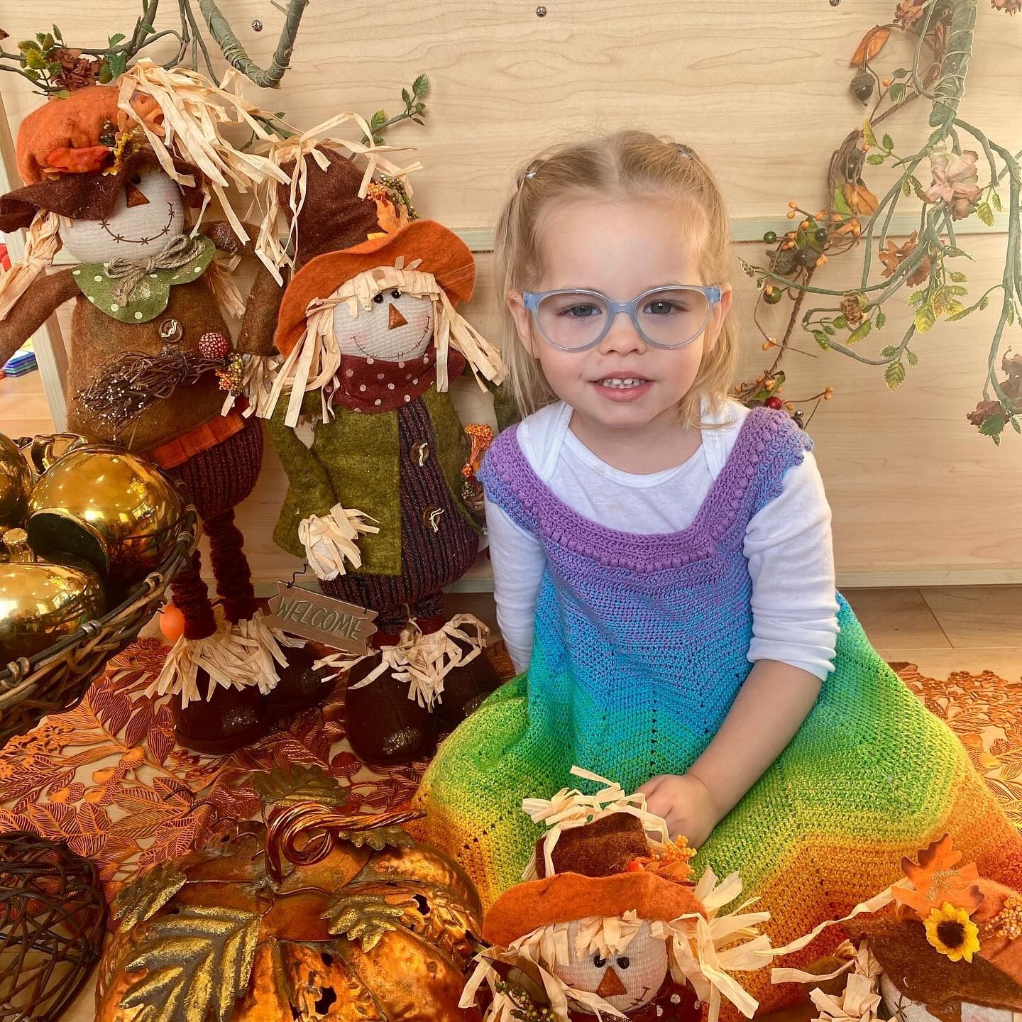 Girl in rainbow dress and glasses, smiling near scarecrows and autumn decor.