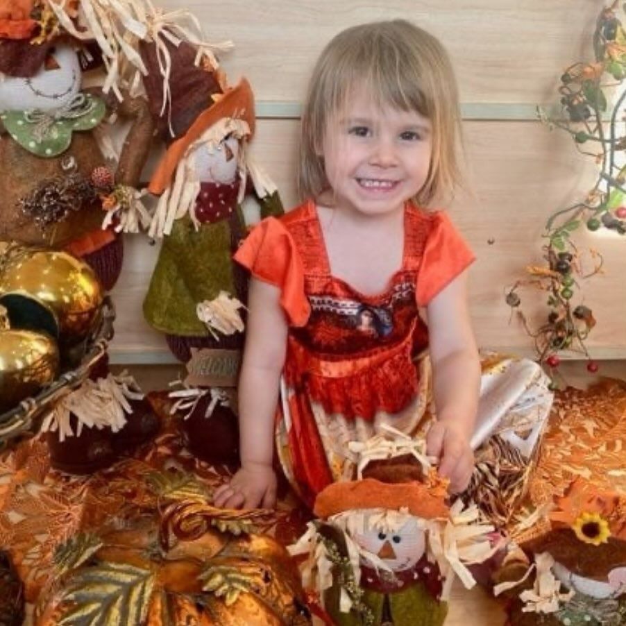 Young child smiles amidst fall decorations, including scarecrows and pumpkins.