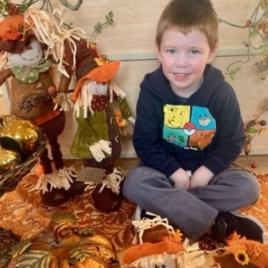 Boy sitting cross-legged smiles in autumn scene with scarecrows, pumpkins, and fall leaves. He wears a blue hoodie and gray pants.