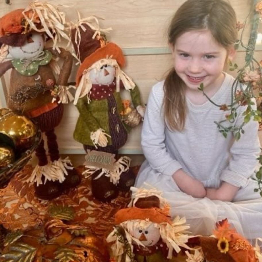 Girl smiles next to fall scarecrow decorations, surrounded by autumn colors.