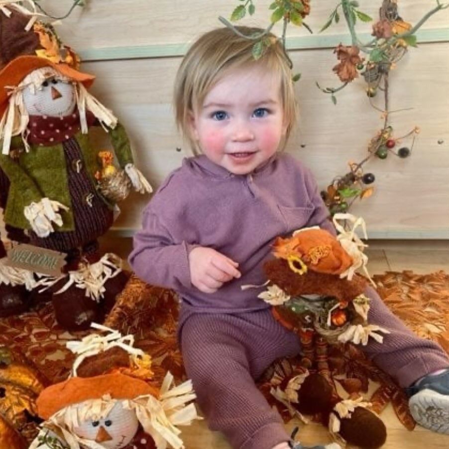 Child in a mauve outfit smiles, holding a scarecrow toy surrounded by fall decor.