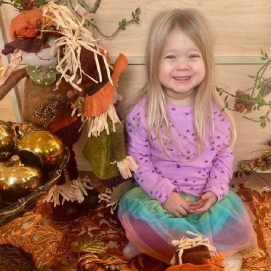 Smiling child with blonde hair wearing a purple shirt and rainbow skirt, sitting near fall decorations and a scarecrow.