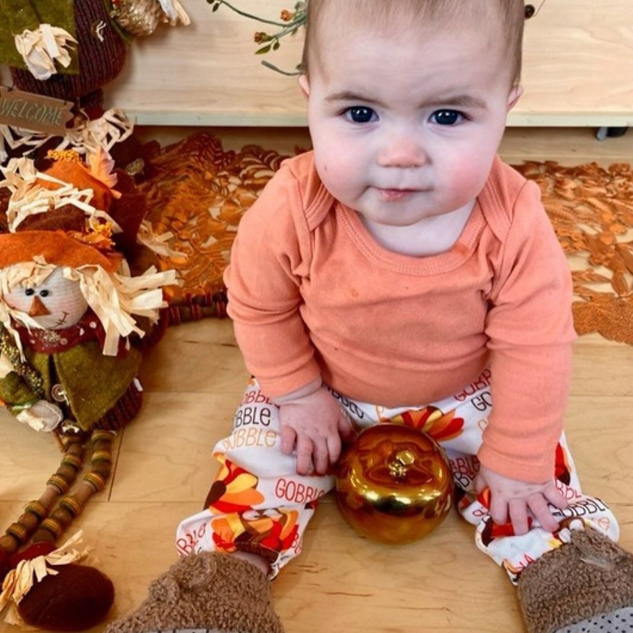 Baby in orange top, fall-themed pants, and sitting near a golden apple and scarecrow decoration.