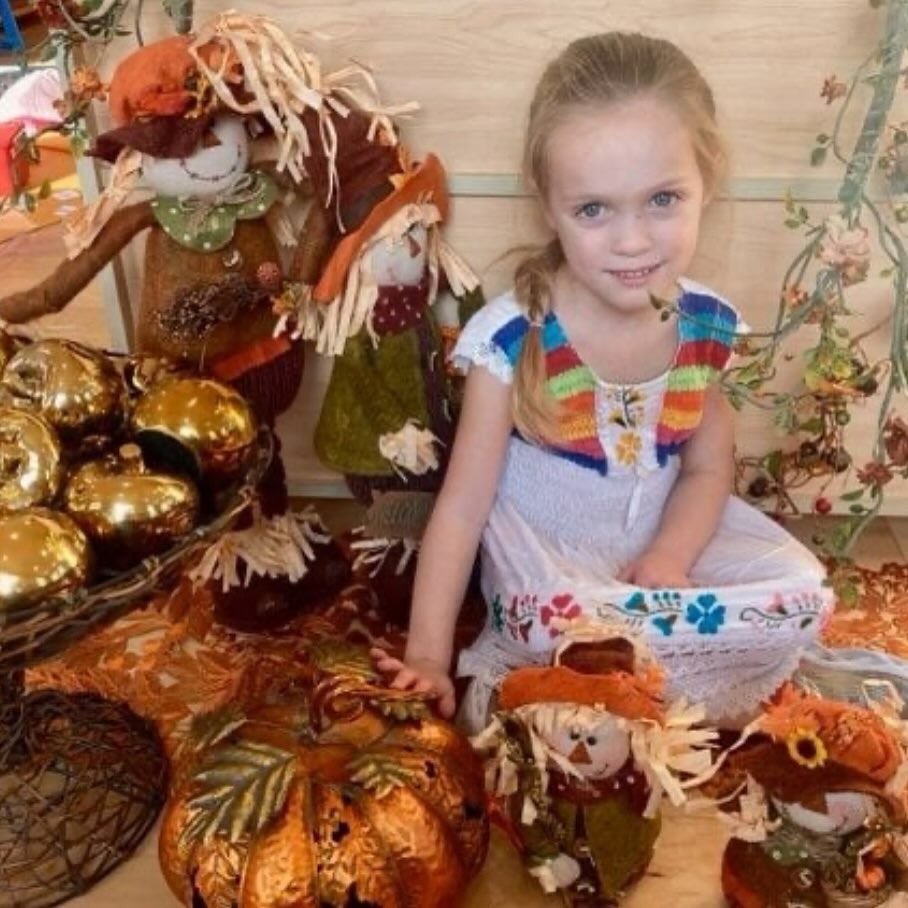 Girl with blonde hair surrounded by fall decorations, including scarecrows and pumpkins.