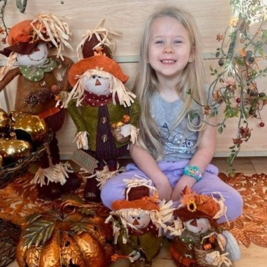 Girl with long blonde hair smiles, surrounded by fall decorations like scarecrows and pumpkins.