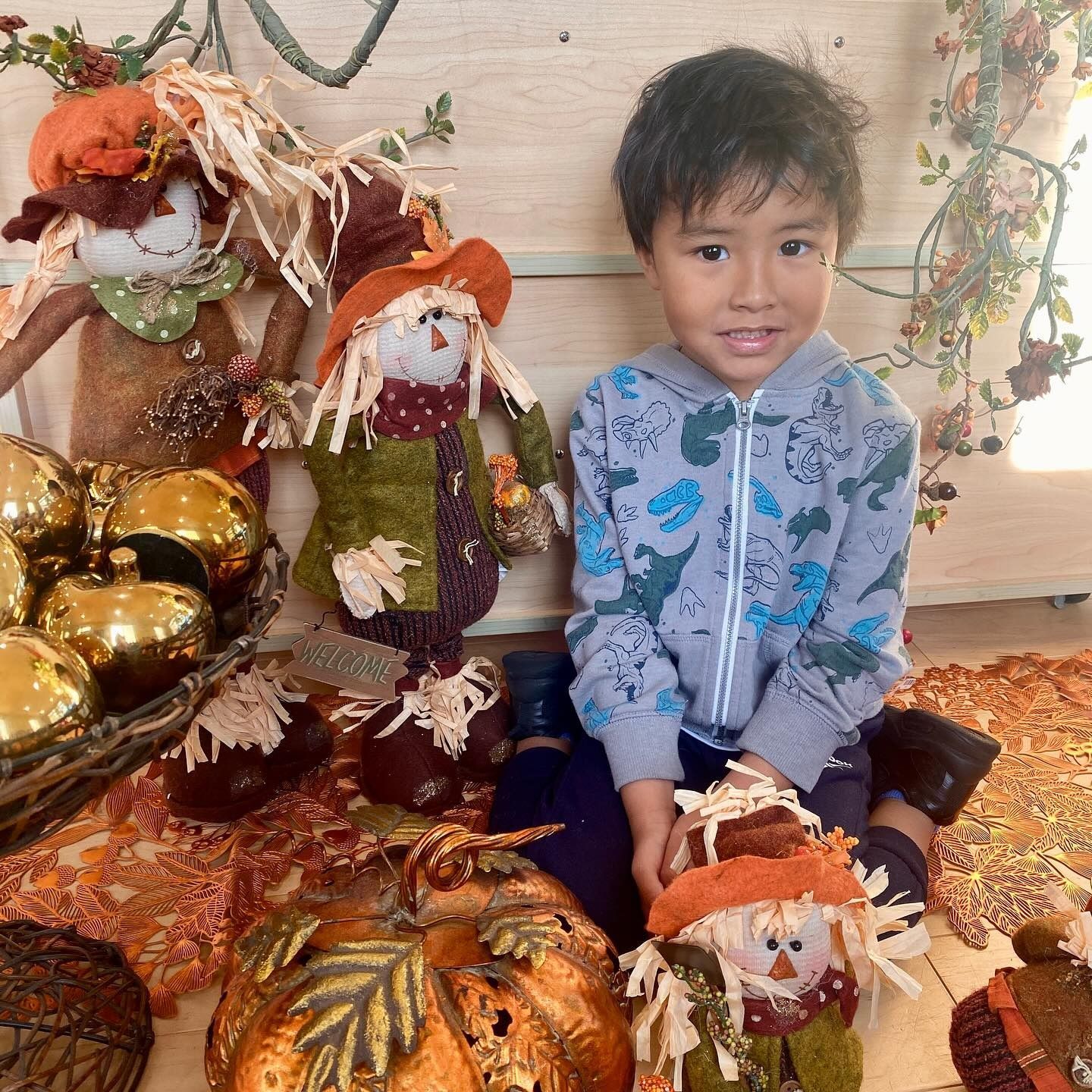 Boy smiles, surrounded by autumn decorations: pumpkins, scarecrows, and golden ornaments.