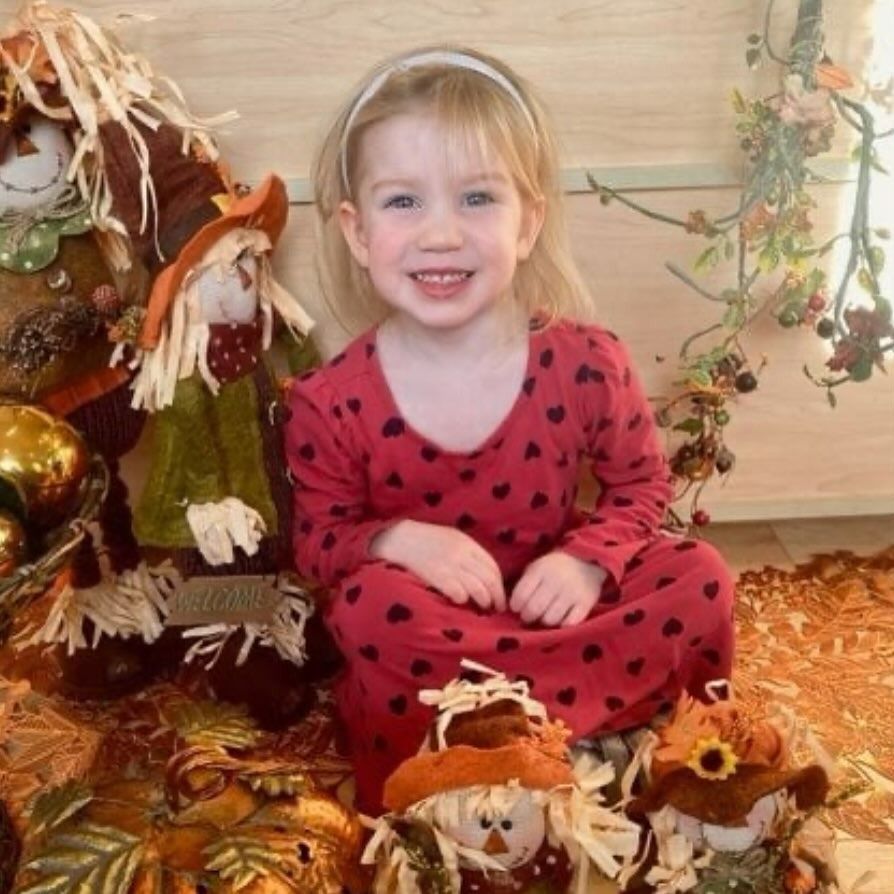 Smiling child in red heart-print dress, sitting amid fall decorations with scarecrows and foliage.