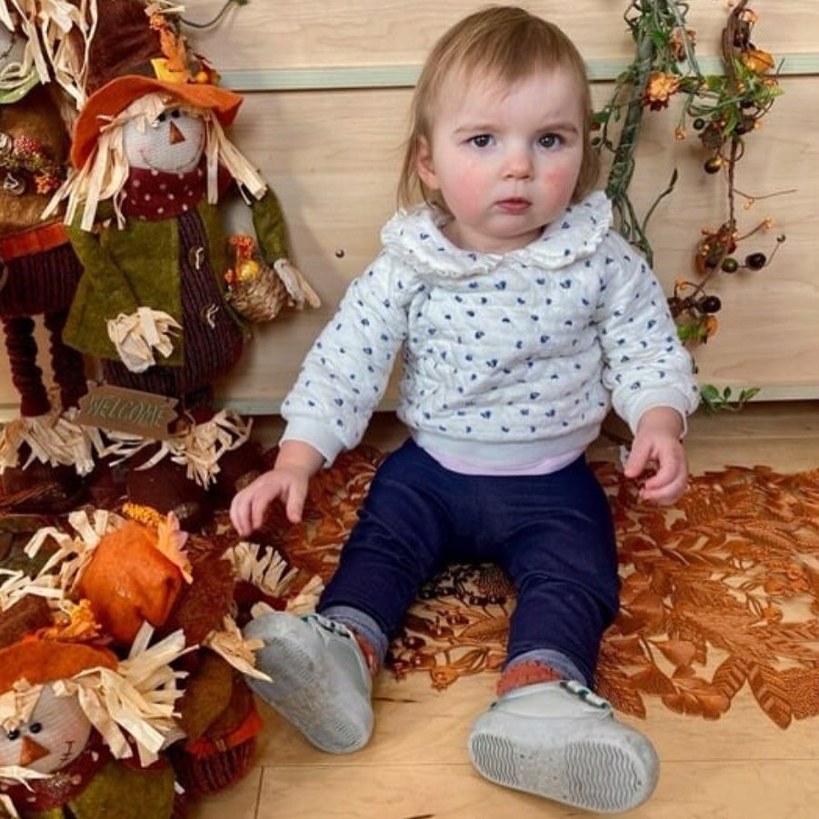 Child seated on fall leaves, wearing blue pants and a sweater with heart details, near a scarecrow and fall decor.