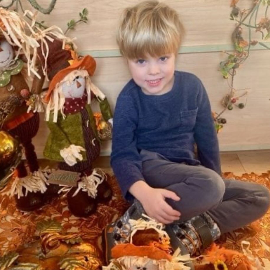 Boy smiles, seated near a scarecrow and fall decorations.