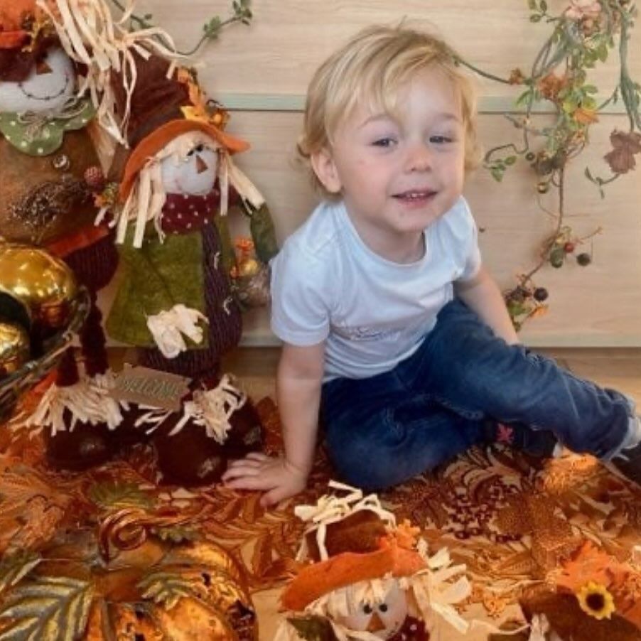 Blonde toddler smiling, seated amidst fall decorations: scarecrows, leaves.