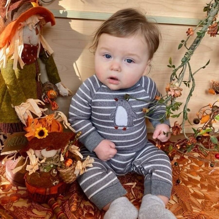 Baby in gray striped onesie with penguin on it, sits amidst fall decorations.