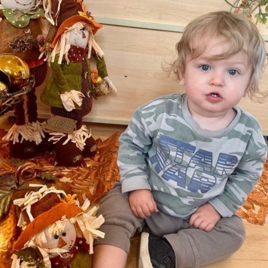 A young child sits, looking at the camera, surrounded by fall decorations.