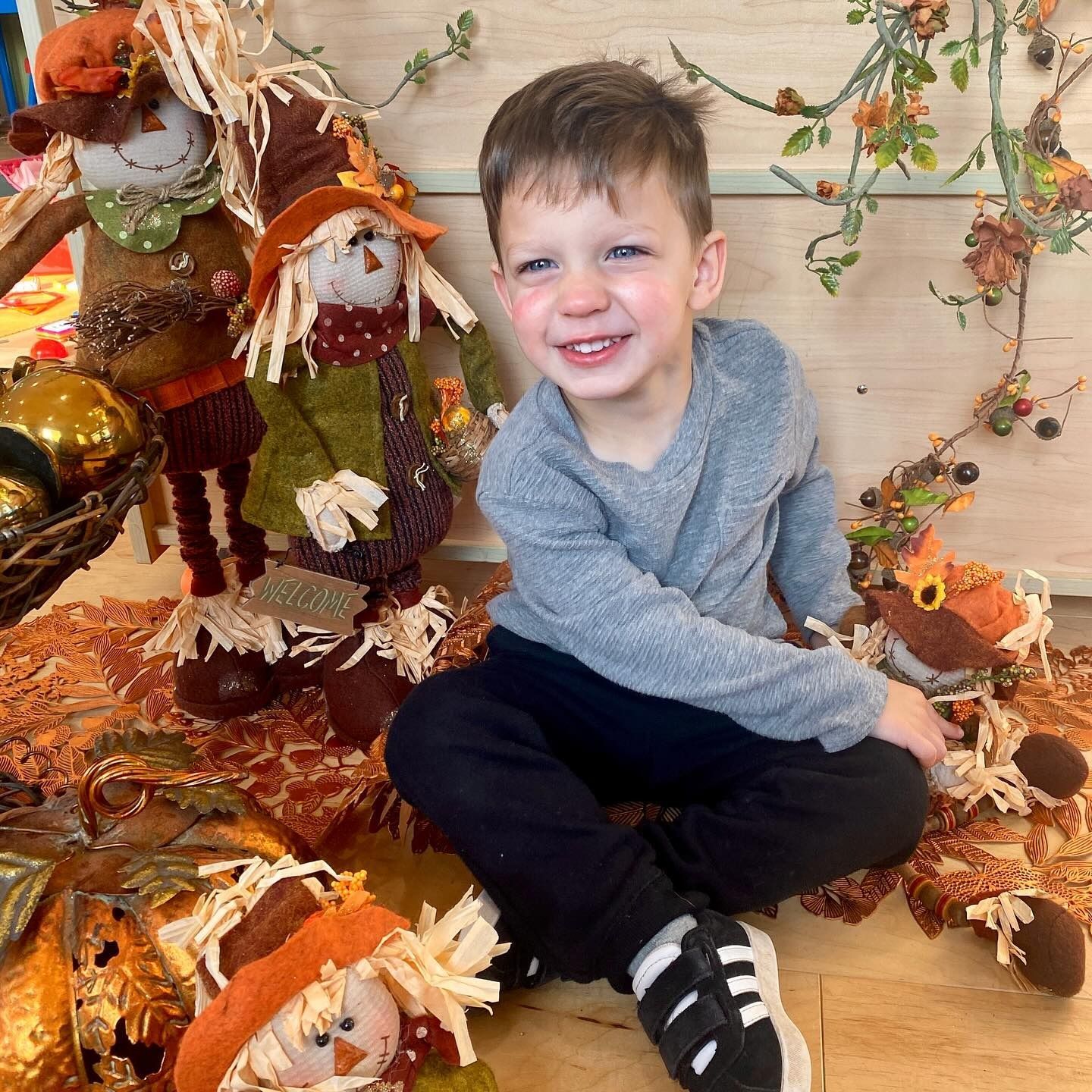 Boy smiling, sitting among fall decorations including scarecrows and leaves.