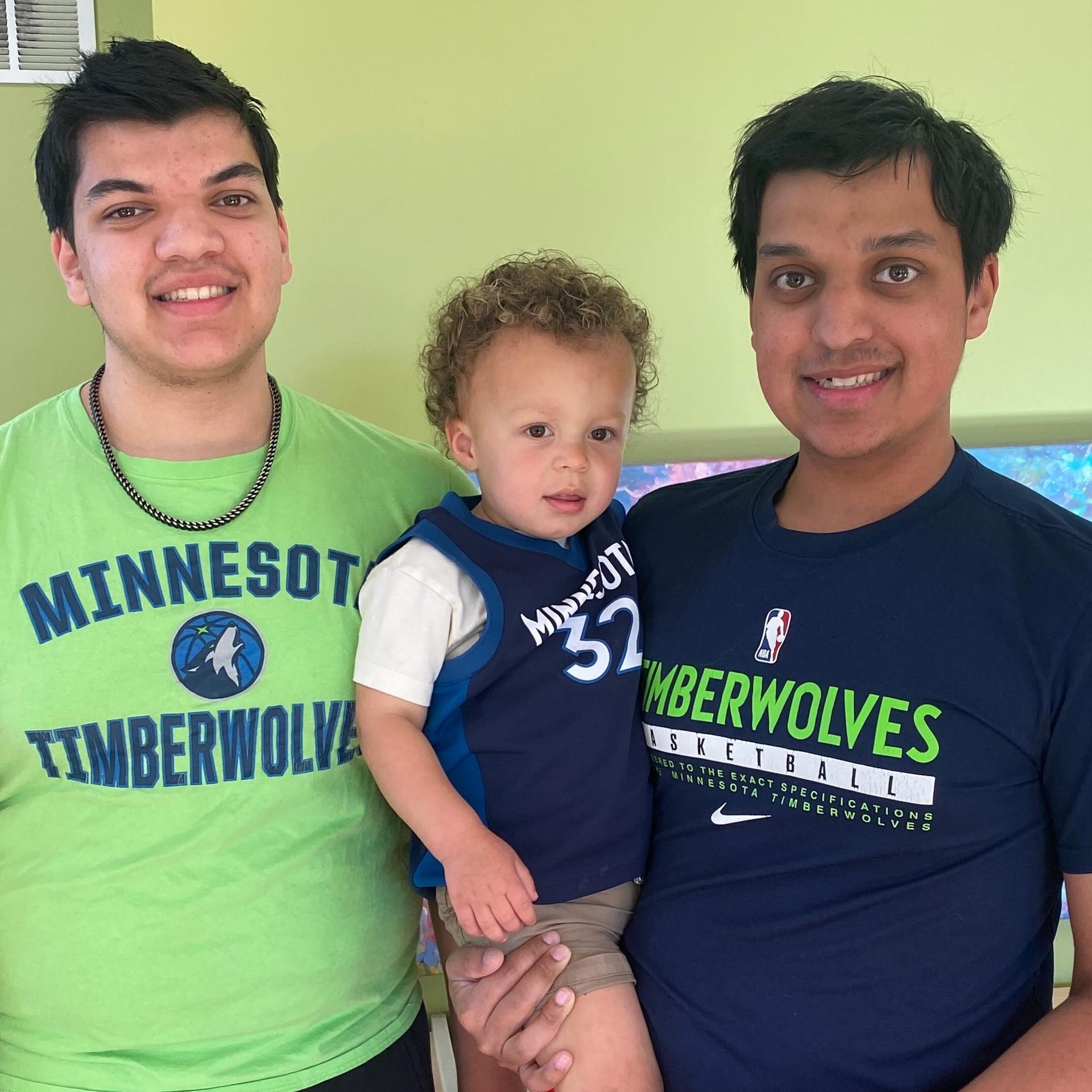 Two men and a toddler pose for a photo. The men wear Minnesota Timberwolves shirts. The toddler has curly hair.
