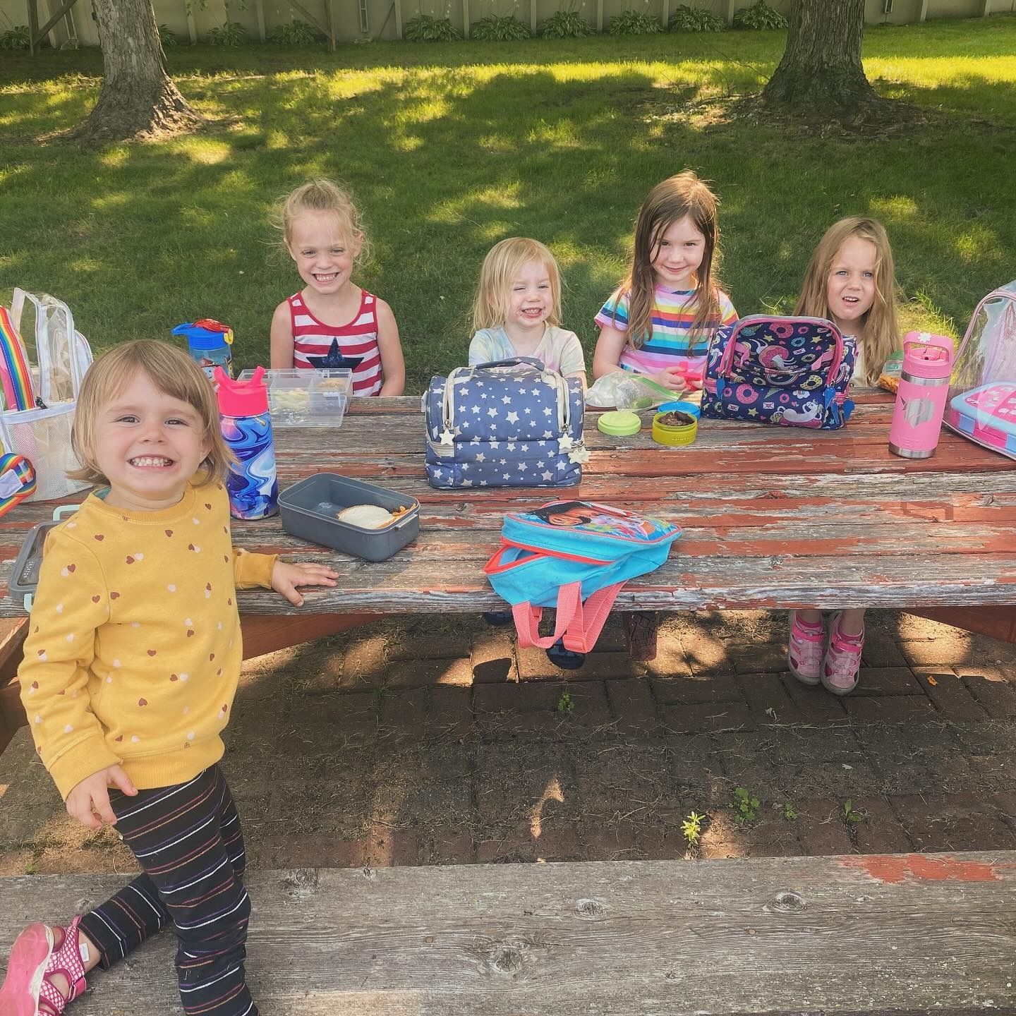 Children at a picnic table, smiling. Lunch bags and food visible. Outdoors in daylight.