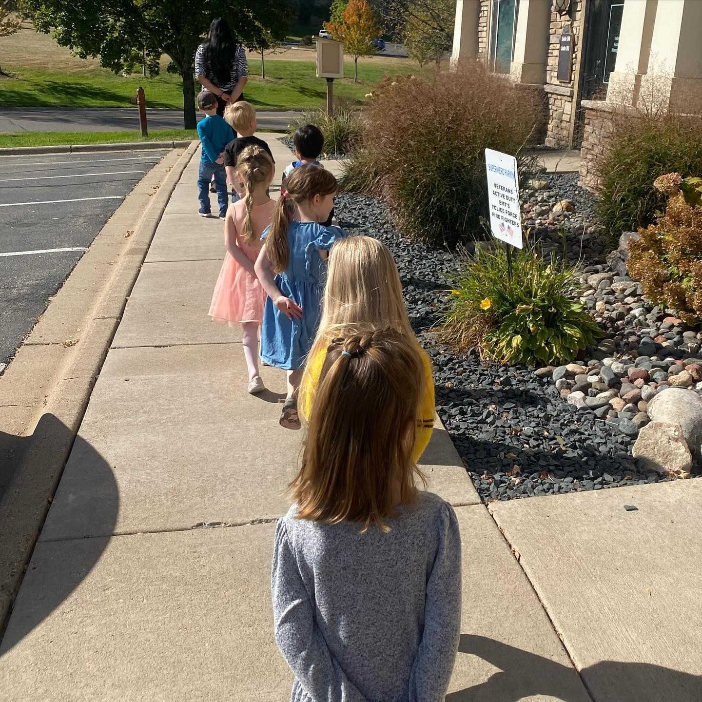 Children lined up on a sidewalk outside a building, following an adult, sunny day.
